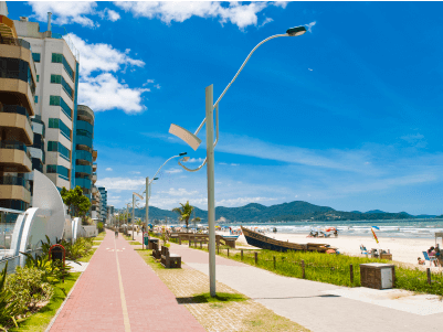 Calçadão de Meia Praia em Itapema - SC, com prédios, postes de iluminação e o mar ao fundo em um dia ensolarado.