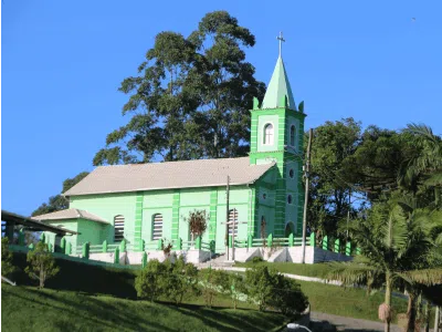 Capela Sertão do Trombudo em Itapema - SC, destacando-se com sua cor verde vibrante, rodeada por árvores e céu azul claro.