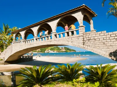 Ponte do Suspiro em Itapema - SC com arcos de pedra e turistas caminhando, emoldurada por palmeiras e com o mar azul ao fundo.